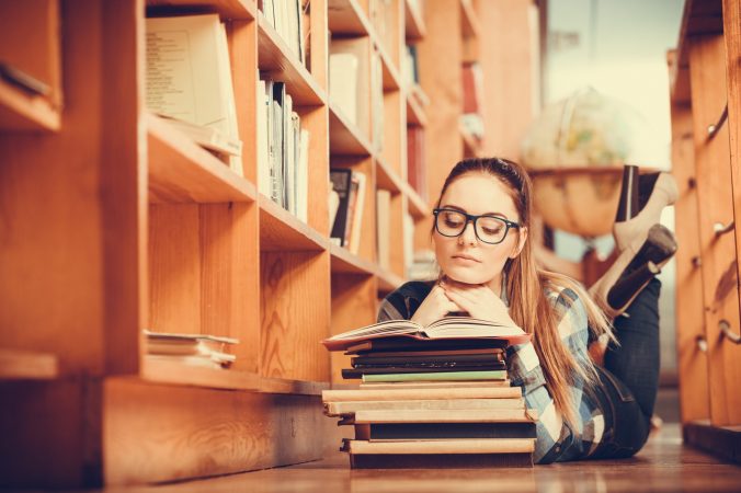 Education school concept. Clever female student girl in glasses lying on floor in college library with stack books reading. Instagram tone